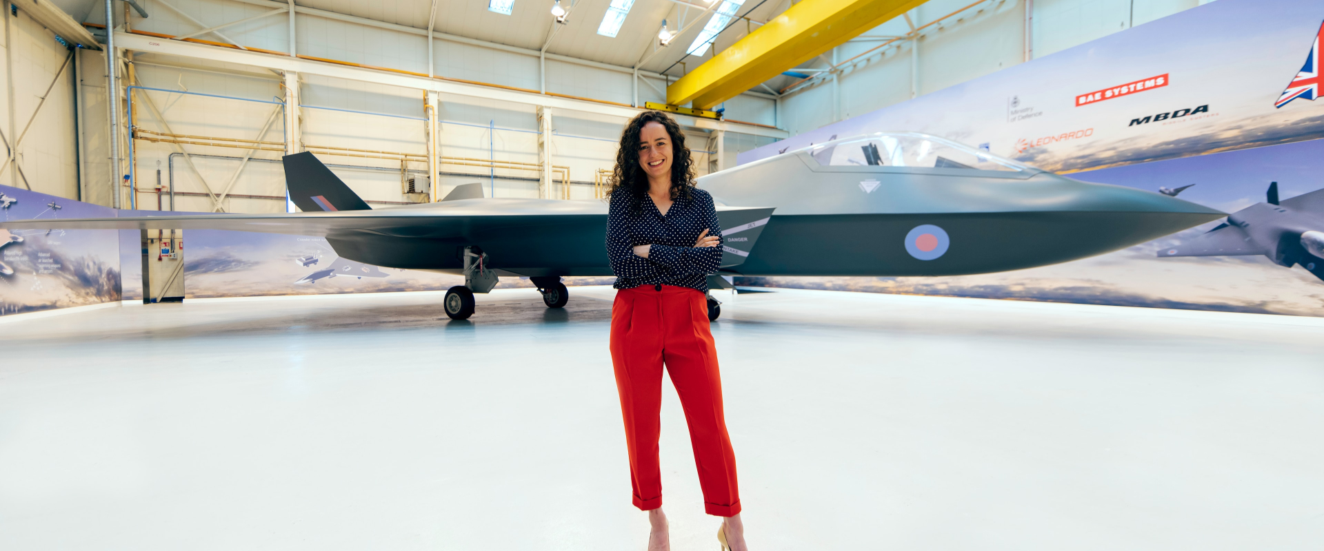 A smiling person standing in front of modern fighter jet in an aircraft hanger.