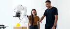 People standing next to a robot arm as it attempts to pick up a yellow ball.