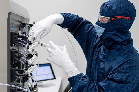 A scientist in protective gear injects fluid into a complex looking machine.