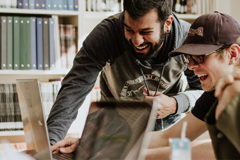 A group of laughing students gathered around a laptop in a library.