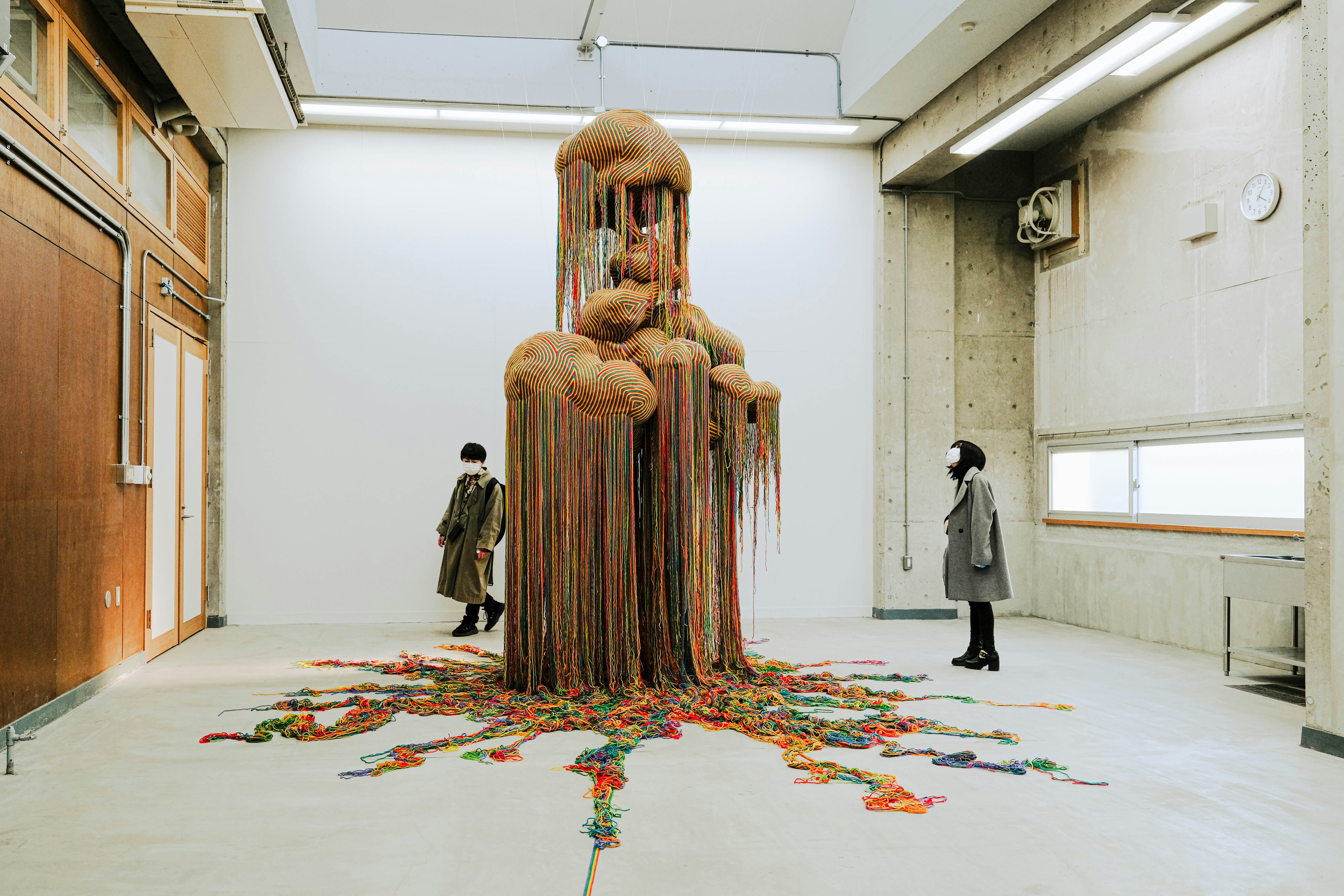 Two people inspecting a large, abstract sculpture of what appears to be multicoloured strands of string hanging down and pooling beneath a fungal structure.